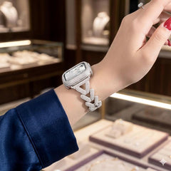 May include: A silver smartwatch with a rectangular face and a crystal-studded band. The watch is worn on a wrist, with a blue sleeve in the background. The watch face is also encrusted with crystals.