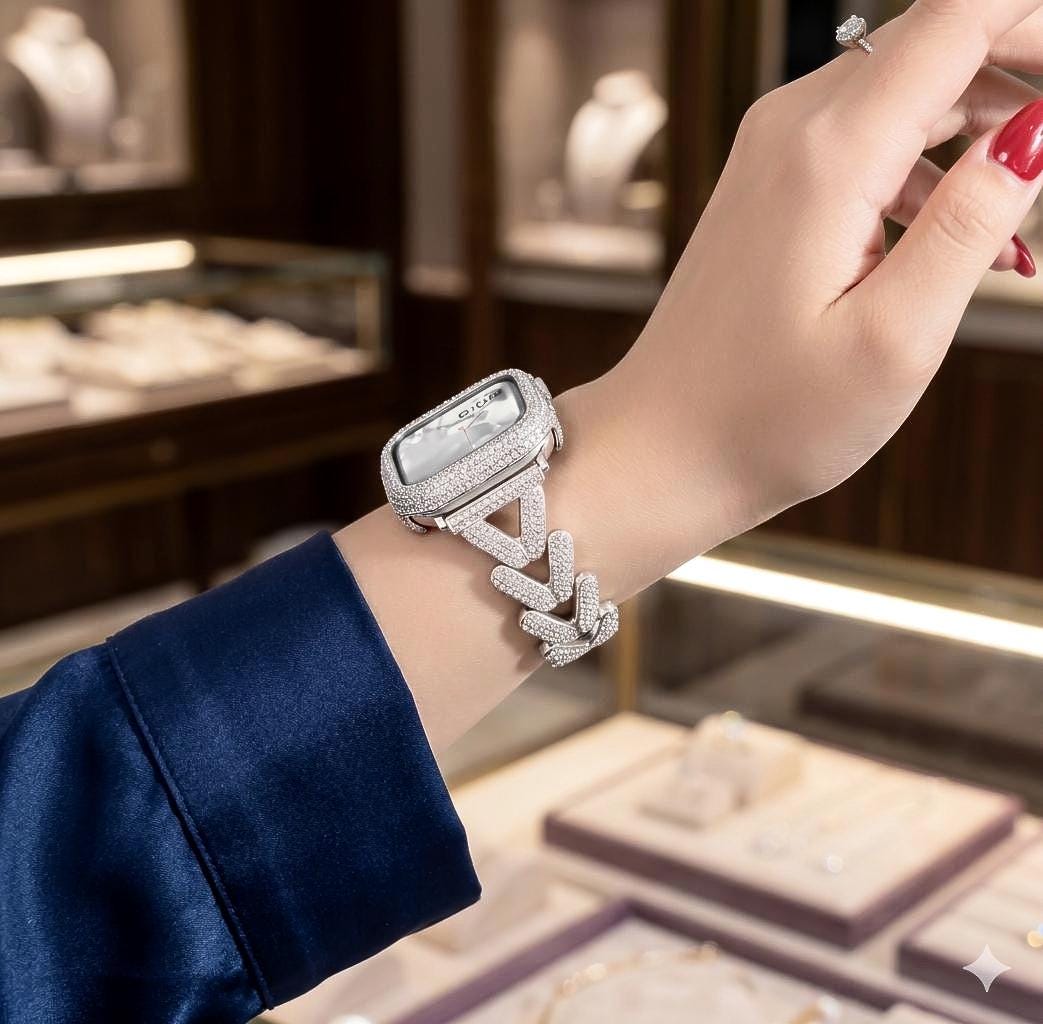 May include: A silver smartwatch with a rectangular face and a crystal-studded band. The watch is worn on a wrist, with a blue sleeve in the background. The watch face is also encrusted with crystals.